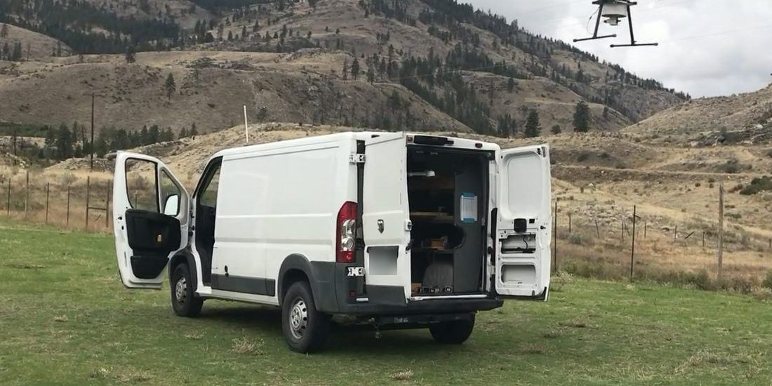 A white M3 van parked in a field with its doors open, the release drone overhead